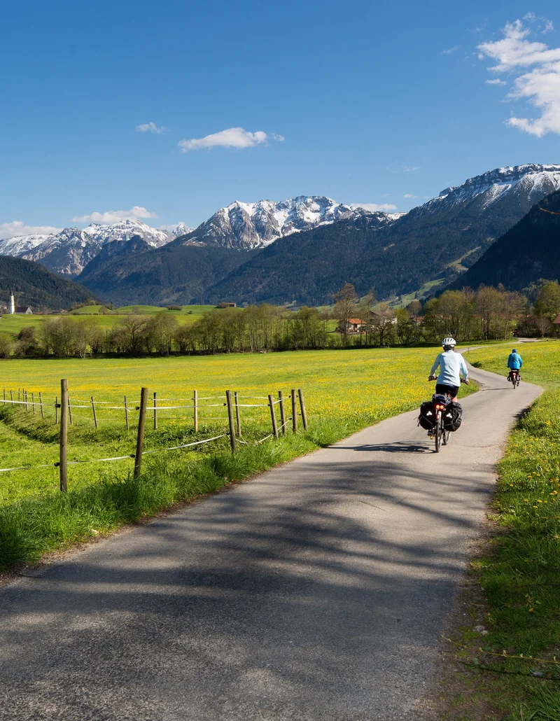 Frühling im Allgäu - bestes Radelwetter Radfahrer auf einem Radweg mit frühlingshaftem Blick auf grüne Wiesen und weiße Berggipfel.