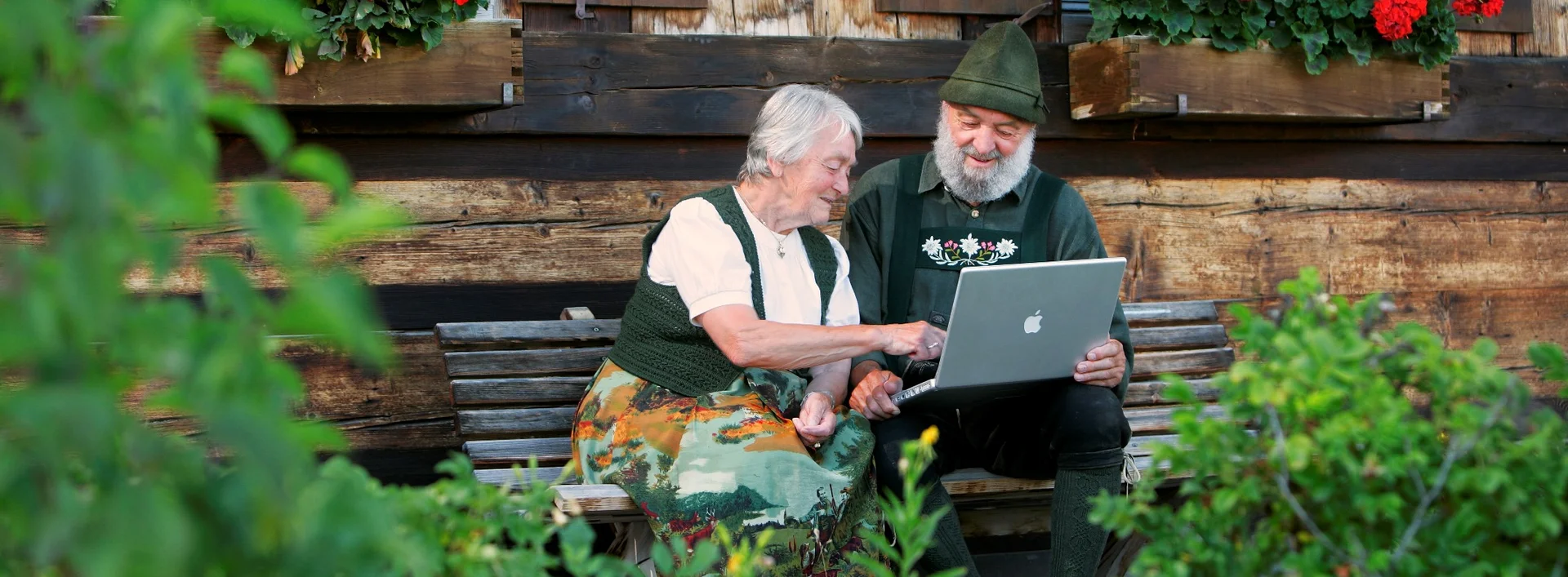 WLAN in Pfronten im Allgäu Zwei Personen in Tracht sitzen auf einer Bank und schauen auf ihren Laptop.