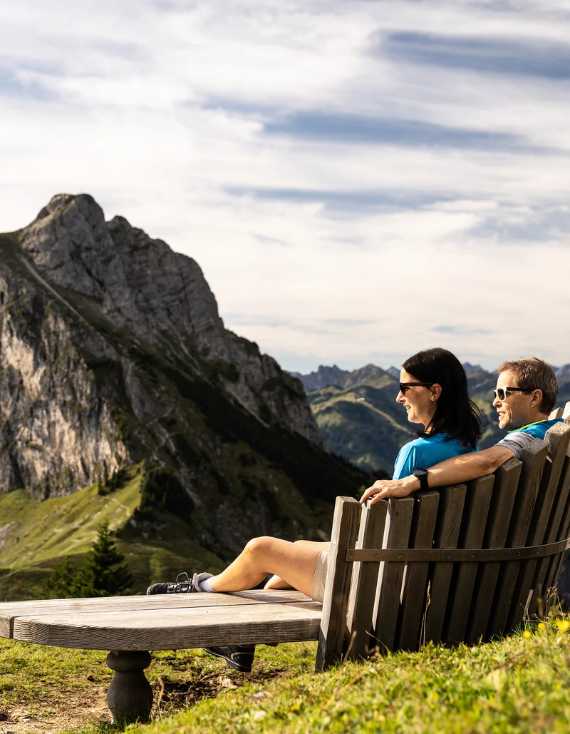 Zwei Wanderer auf einer Sitzbank mit Blick auf die beeindruckende Bergkulisse.