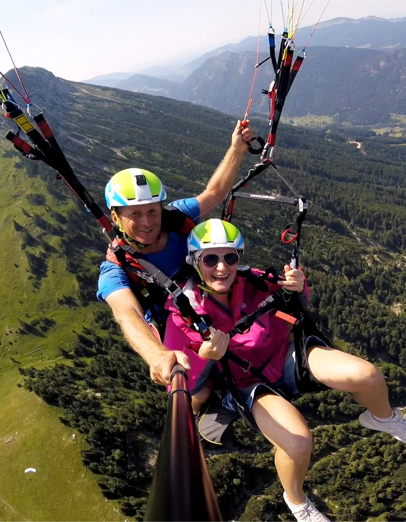 Foto von FlyTeam Ein Tandemflug mit dem Gleitschirm bei Sonnenschein und einer Aussicht auf eine schöne Berglandschaft.