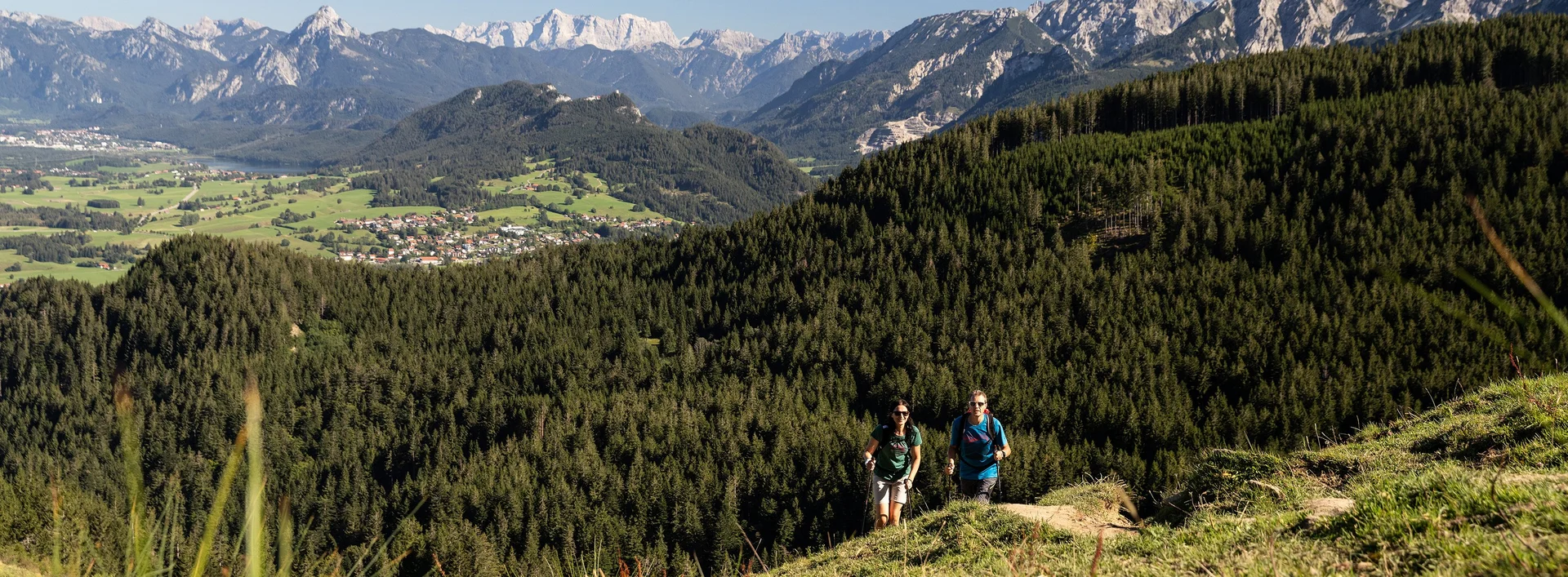 Zwei Wanderer auf einem schmalen Pfad auf dem Berg inmitten grüner Wiesen und einer beeindruckenden Bergkulisse.