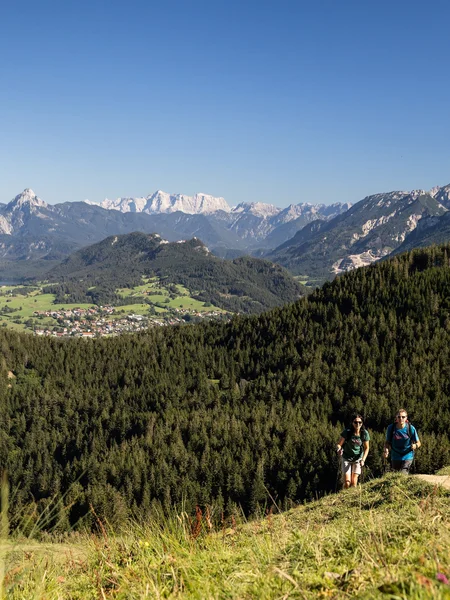 Zwei Wanderer auf einem schmalen Pfad auf dem Berg inmitten grüner Wiesen und einer beeindruckenden Bergkulisse.