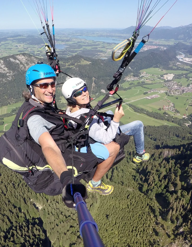 Foto von FlyTeam Ein Tandemflug mit dem Gleitschirm bei Sonnenschein und einer Aussicht auf eine schöne Berglandschaft.