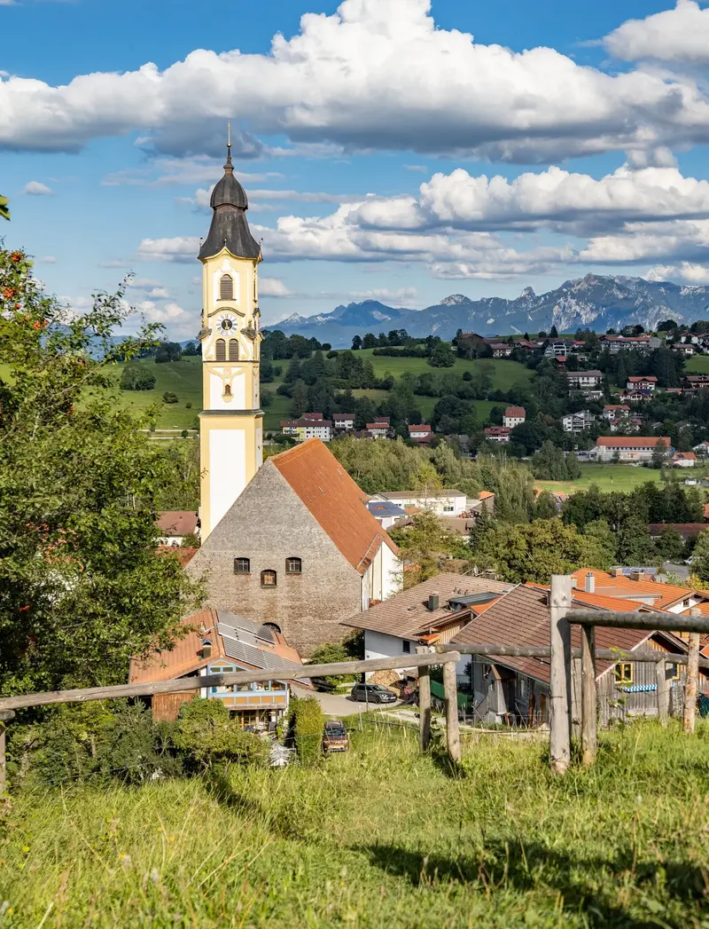 Blick vom Aussichtspunkt "Hörnle" auf die Pfrontener Kirche und die dahinterliegende Berglandschaft.