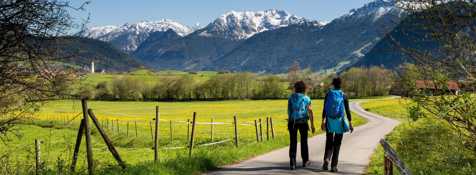 Zwei Wanderer auf einem schönen Weg durch das frühlingshafte Pfrontener Tal mit Blick auf die weißen Berggipfel.