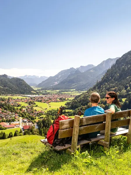 Zwei Wanderer sitzen auf einer Bank auf dem Edelsberg und genießen die Aussicht über Pfronten und die dahinterliegende Berglandschaft.