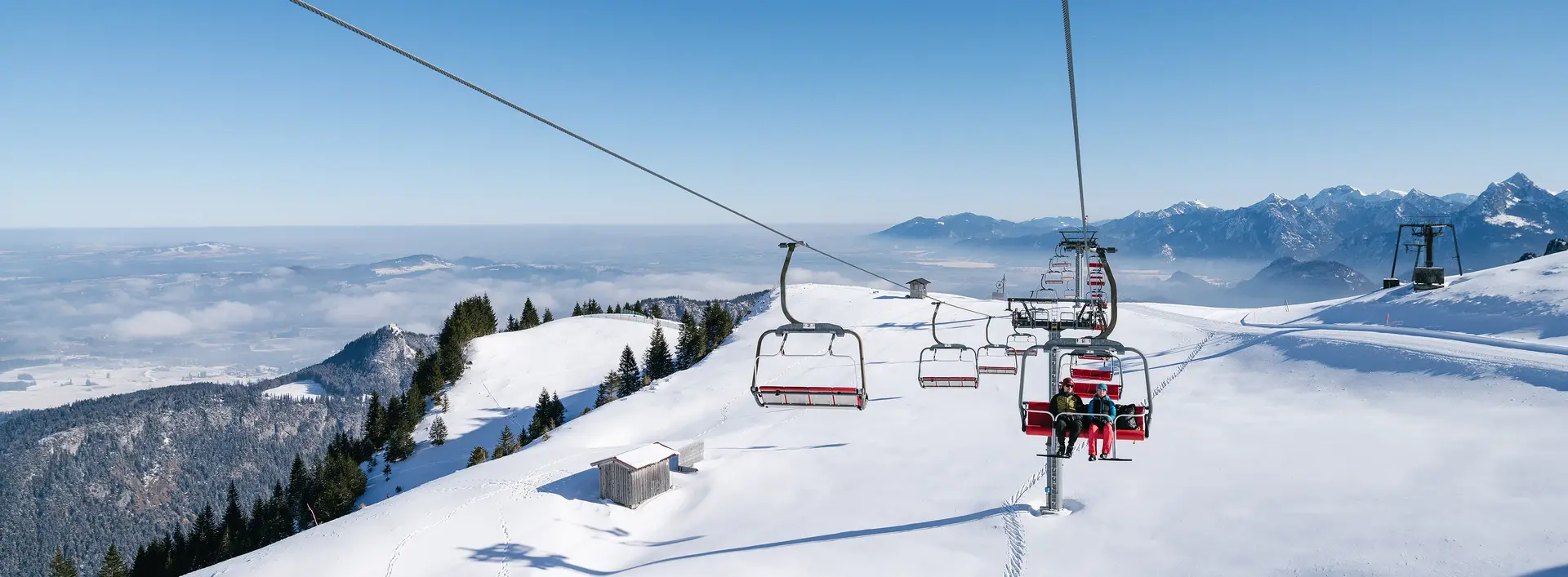 Blick von der Sesselbahn am Breitenberg über eine idyllisch verschneite Winterlandschaft mit den Allgäuer Bergen im Hintergrund.