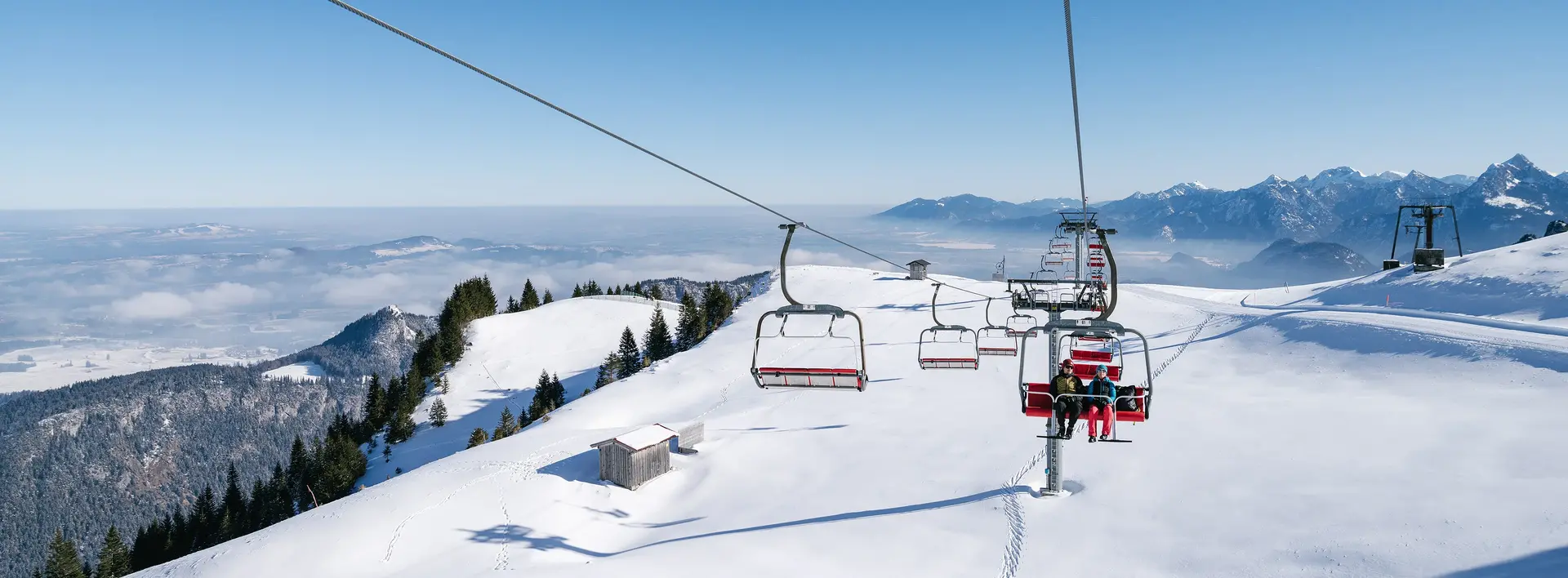 Blick von der Sesselbahn am Breitenberg über eine idyllisch verschneite Winterlandschaft mit den Allgäuer Bergen im Hintergrund.