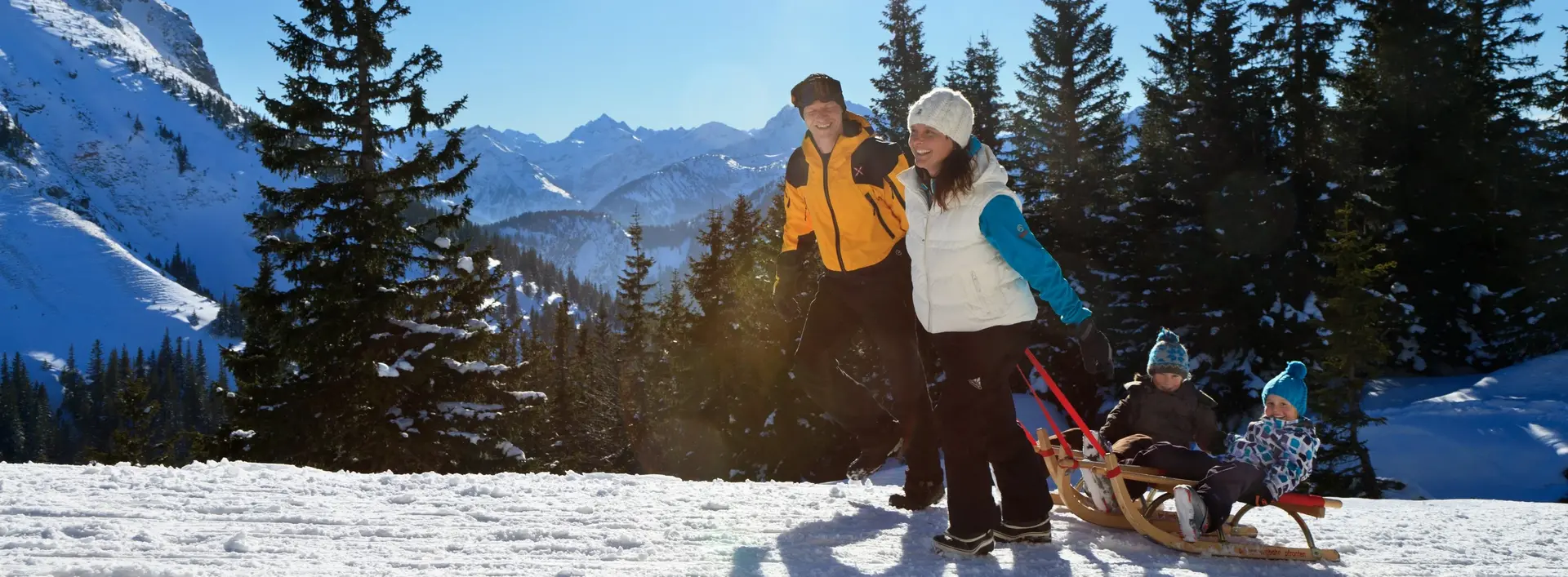 Zwei Kinder sitzen auf einem Schlitten und werden von ihren Eltern an einem schneebedeckten Winterweg gezogen.