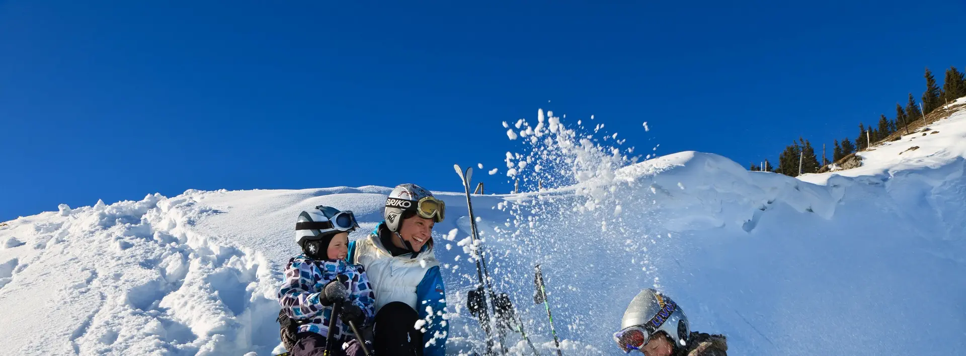 Eine Skifahrerfamilie sitzt bei sonnigem Wetter im Schnee und hat Spaß.